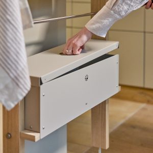 Mano che apre il coperchio a ribalta di un contenitore WoodUpp montato su struttura in legno chiaro: scatola bianca metallica con foro per presa, finitura opaca, viti a vista e logo traforato sul fronte. Prodotto WoodUpp per organizzazione armadio o porta biancheria.