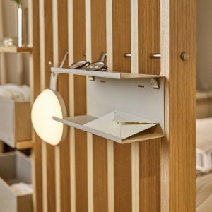 Pannello divisorio in legno WoodUpp con listelli verticali chiari; fissato uno scaffaletto a parete WoodUpp in metallo beige a doppio ripiano che contiene un paio di occhiali e una busta, agganciato a ganci metallici. Presente anche una lampada rotonda sospesa e bullone decorativo.