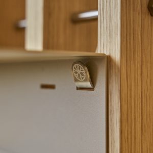 Primo piano di un sistema WoodUpp: pannello modulare in rovere chiaro con doghe verticali e un supporto/gancio in metallo brunito fissato a una piastra grigia. Il gancio presenta un decoro circolare inciso a raggiera, parte degli accessori metallici per scaffalature WoodUpp.