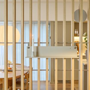 Paramento a lamelle di legno con separé interno; fissata una mensola WoodUpp in metallo color sabbia con piccolo portacandela integrato che sostiene una candela bianca accesa; sullo sfondo tavolo da pranzo, lampada rotonda e cucina, minimalista e luminosa con tende chiare.