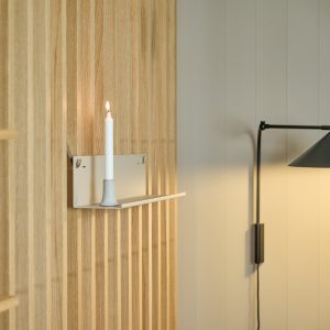 Pannello a doghe in legno chiaro WoodUpp riveste la parete; una mensolina metallica bianca WoodUpp sottile è fissata al pannello e regge un portacandela in ceramica con candela bianca accesa. A destra appare una lampada da parete nera regolabile, scena minimalista.