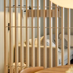 Parete divisoria a lamelle verticali in rovere chiaro WoodUpp, con collegamenti e staffe metalliche visibili, che separa gli ambienti. Sullo sfondo un divano beige con cuscini e un cesto; vista ravvicinata delle doghe in legno levigato e della struttura modulare WoodUpp.