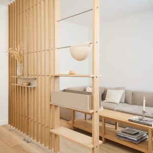 Paravento modulare WoodUpp in rovere chiaro con listelli verticali: mensole regolabili, ripiano sospeso con vaso e ciotola, cassettino contenitore in feltro beige estraibile e lucina a mezza sfera. Sistema divisorio minimal che separa il living con divano, tavolino e libri decorativi e funzionale.