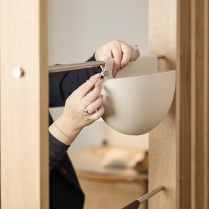 Mani che fissano a una struttura in legno chiaro un vaso sospeso semisferico beige di WoodUpp, agganciato a un'asta metallica tramite un piccolo gancio clip metallico. In primo piano mani curate con anelli e braccialetto evidenziano il design moderno e minimal del prodotto.