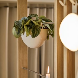 Pannello modulare in legno WoodUpp con listelli verticali e aste metalliche, supporto con vaso sospeso bianco WoodUpp a semisfera contenente una pianta dalle foglie verdi; lampada rotonda diffusa e una candela accesa in primo piano, atmosfera calda e naturale elegante.