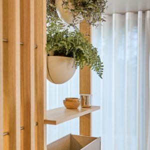 Struttura modulare in legno WoodUpp dalla finitura naturale: parete divisoria con montanti verticali, mensola a sbalzo e vasetti sospesi semicircolari bianchi contenenti felci rigogliose, più due ciotoline in legno e un piccolo vaso metallico sul ripiano. Atmosfera luminosa con tende chiare sullo sfondo.