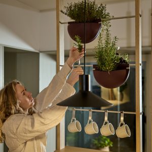 Mobile modulare WoodUpp in legno con due vasi sospesi semisferici bordeaux contenenti erbe aromatiche, barra metallica con ganci e quattro tazze bianche appese; una donna sistema le piante, davanti a una lampada nera a sospensione che domina la scena, calda.