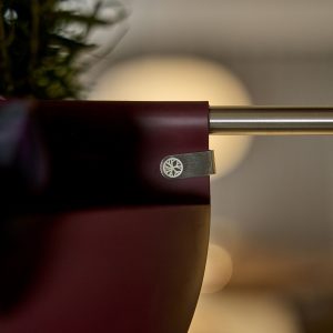 Primo piano di un vaso appeso WoodUpp color bordeaux fissato a una bacchetta metallica dorata tramite una clip in acciaio con logo circolare a raggiera di WoodUpp; si intravede una pianta nella parte superiore e uno sfondo sfocato con luci calde.