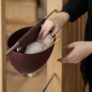 Mano che prende un gomitolo da un contenitore sospeso WoodUpp color borgogna, montato su una parete in legno con barra metallica: contenitore per lavori a maglia che contiene filo chiaro e ferri di legno, abbinato al sistema di supporto modulare e versatile WoodUpp.