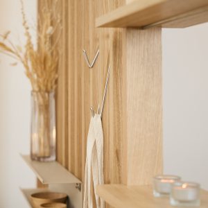 Pannello WoodUpp in legno a listelli con mensole sottili e ganci metallici a V e a doppia punta WoodUpp; vaso di vetro con fiori secchi, ciotole di legno, due candele in portacandele di vetro e una stoffa chiara appesa a un gancio