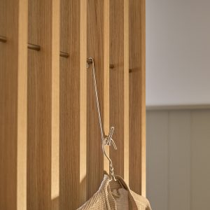 Pannello verticale WoodUpp in listelli di rovere chiaro con piccoli perni in ottone, su cui è appeso un gancio metallico sottile che sostiene un capo leggero a trama; immagine ravvicinata evidenzia venatura del legno e dettaglio dei ganci metallici WoodUpp.