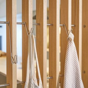Schermo divisorio WoodUpp in listelli verticali di rovere chiaro, con barre orizzontali metalliche e ganci WoodUpp in acciaio inox; due ganci sostengono una borsa tessile chiara e un canovaccio a righe. Illuminazione morbida sullo sfondo, dettagli materici e finiture naturali.