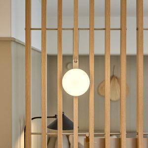 Divisorio a listelli in rovere WoodUpp con barra metallica orizzontale e gancio WoodUpp che sostiene una lampada tonda sospesa dal design moderno; sullo sfondo si intravede una lampada da tavolo nera e dettagli di un ambiente domestico accogliente dallo stile minimalista e colori neutri.