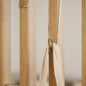 Primo piano di un pannello WoodUpp: listelli verticali in rovere chiaro con ganci sottili in acciaio inox fissati, utilizzati come appendiabiti. Si vede una tracolla di tessuto beige appesa a uno dei ganci, dettaglio che enfatizza stile minimal e finitura naturale.
