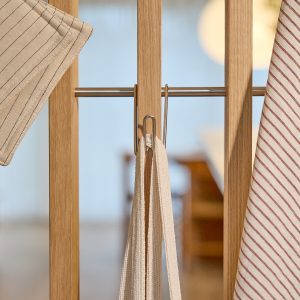 Pannello divisorio WoodUpp in rovere chiaro con barra appendiabiti in acciaio inox e due ganci metallici WoodUpp che sorreggono strofinacci e guanto da cucina in cotone a righe beige. Dettaglio minimal, finiture naturali e linee pulite, atmosfera domestica calda e praticità quotidiana.