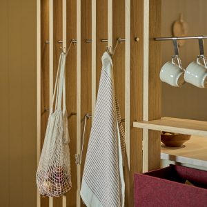 Dettaglio di una parete divisoria modulare WoodUpp in listelli di legno chiaro con asta metallica, ganci in acciaio e mensola integrata WoodUpp; appendono una borsa a rete con cipolle, un canovaccio a righe e tazze smaltate, accanto a un contenitore in feltro bordeaux.