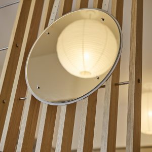 Pannello divisorio WoodUpp in doghe verticali di rovere chiaro, con aste metalliche orizzontali, completo di specchio tondo montato su distanziali cromati; nello specchio si riflette una grande sospensione di carta bianca. Design lineare, materiali naturali e dettagli metallici visibili, ideale per separare ambienti e valorizzare la luce naturale.