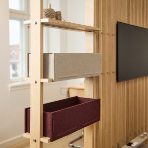 Immagine WoodUpp che mostra un divisorio in legno chiaro con mensole modulari e due contenitori in feltro WoodUpp: uno grigio chiaro e uno bordeaux, inseriti su supporti a guida. Sul ripiano superiore spago avvolto; sullo sfondo parete dogata in legno e televisore nero.