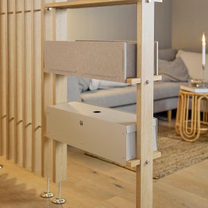 Divisorio modulare WoodUpp in rovere chiaro con montanti verticali, due accessori WoodUpp: un contenitore in feltro marrone inserito su mensola e una scatola porta‑cavi metallica beige con foro superiore e prese d'aria, fissati con bulloni e supporti visibili in un interno living caldo.