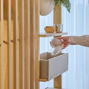 Mobile modulare WoodUpp in rovere con pannelli a listelli verticali e mensole; si vedono un vaso appeso bianco con pianta, ciotole in legno e un bicchiere metallico sulla mensola, e un cassetto removibile grigio in feltro con una mano che ripone gomitoli di lana.