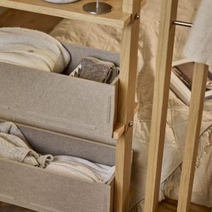 Mobile modulare WoodUpp in legno di rovere chiaro con due contenitori portaoggetti in tessuto grigio-beige inseriti come cassetti, ripiani sottili e fissaggi metallici; nei box si vedono maglioni e calzini piegati, posizionato accanto al letto con copriletto naturale, dallo stile minimal e tonalità neutre.