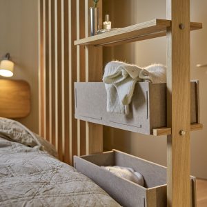 Divisorio modulare WoodUpp in rovere con montanti verticali e ripiani, dotato di cassette portaoggetti in feltro beige WoodUpp. Si vede un maglione chiaro ripiegato nel contenitore superiore, un secondo cassettino vuoto sotto e mensola superiore con vaso e profumo, accanto al letto.