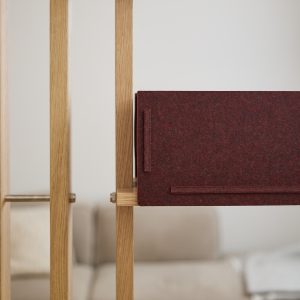 Struttura modulare in rovere chiaro WoodUpp con montanti verticali e connettori in ottone, affiancata da un pannello divisorio in feltro bordeaux WoodUpp, rettangolare con ganci in feltro, che funge da schermo decorativo e fonoassorbente su una mensola interna dal design minimalista e raffinato.
