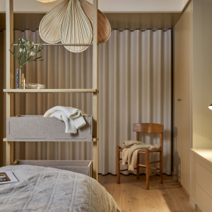 L'immagine mostra una camera da letto caratterizzata da un divisorio a scaffale in legno chiaro di WoodUpp. Questo mobile funzionale WoodUpp integra ripiani aperti e pratici cassetti in feltro grigio, contenenti un maglione, fungendo da separatore d'ambiente elegante e soluzione di archiviazione.