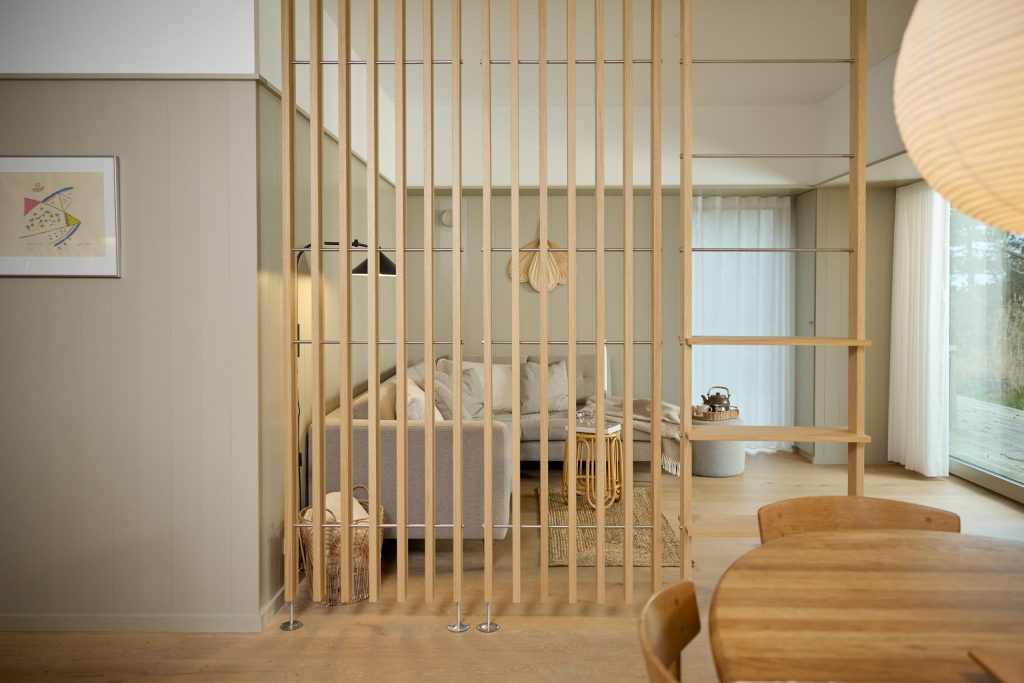 Interno luminoso con divisorio a listelli in rovere firmato WoodUpp che separa soggiorno e zona pranzo; sul lato destro mensole modulari in legno WoodUpp integrate. Si intravedono divano grigio, tavolo da pranzo in legno chiaro e ampie finestre con tende.