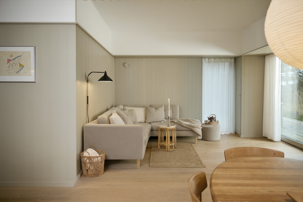 Un soggiorno luminoso e moderno con pareti rivestite dai pannelli da parete WoodUpp in una tonalità beige chiaro. I pannelli verticali con scanalature sottili definiscono lo spazio, creando un'atmosfera calda e accogliente attorno al divano angolare e ai mobili in legno.