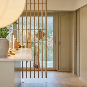 Interno cucina elegante con divisorio verticale WoodUpp a listelli di legno chiaro fissato da pavimento a soffitto, con barre metalliche orizzontali e ganci WoodUpp che reggono strofinaccio e shopper a rete. Sul bancone candela, vaso e ciotole, vista vetro piovoso sul giardino.