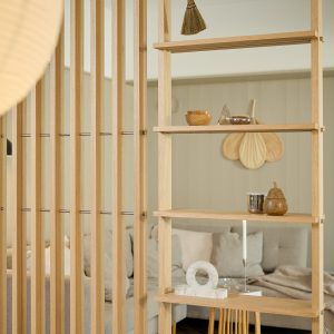 Separé e scaffalatura WoodUpp in rovere chiaro: pannello a listelli verticali con ripiani aperti integrati. Sui piani oggetti decorativi (ciotola in legno, scultura in vetro, tazza metallica, pera in legno, anello in marmo e candela). Sullo sfondo divano e cuscini beige.