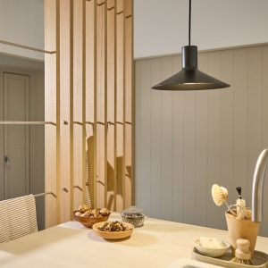Divisorio verticale WoodUpp in listelli di rovere chiaro con aste metalliche e ganci integrati, usato come separazione in cucina; sul piano di lavoro ciotole in legno, vasetto di vetro e utensili, spazzola vicino al lavello e una lampada a sospensione nera sopra.