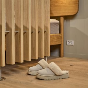 Dettaglio interno WoodUpp: pannello a listelli verticali in rovere chiaro con venature naturali ben evidenti e gambe metalliche sottili, testiera di letto in legno massello e pavimento coordinato. In primo piano ciabatte imbottite beige su parquet, atmosfera calda e naturale.
