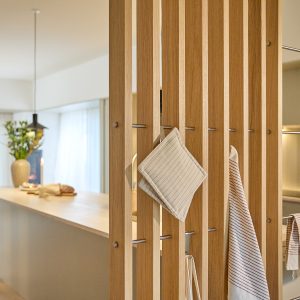 Parete divisoria a listelli in rovere WoodUpp con sottili barre appendino in acciaio WoodUpp, su cui sono appese una presina quadrata beige a righe e uno strofinaccio rigato. Sullo sfondo isola cucina chiara con vaso, candela e pane, atmosfera calda e minimal.