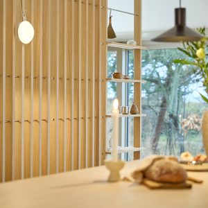 Parete divisoria a listelli verticali in rovere chiaro WoodUpp, finitura naturale, con ganci metallici integrati e ripiani modulari regolabili WoodUpp che ospitano piccoli oggetti in ceramica e legno. In primo piano tavolo con candela accesa e pane; finestra e giardino sullo sfondo.