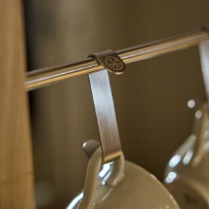 Primo piano su una barra metallica con clip metallica WoodUpp incastrata, che sostiene un gancio appenditazza WoodUpp in acciaio inox. Al gancio è appesa una tazza bianca in ceramica; sullo sfondo si intravede un'altra tazza sfocata, montate su struttura in legno.