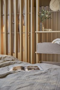 Immagine mostra un ambiente letto con parete separé a listelli di rovere WoodUpp con barre metalliche, scaffale modulare WoodUpp con cassetti in feltro grigio, vaso con fiori, piattino in vetro e trapunta beige sul letto con libro aperto e occhiali.