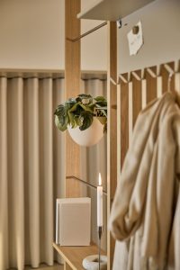 Divisorio modulare WoodUpp dallo stile pulito ed elegante in rovere con mensole sottili, ganci appendiabiti e sbarre metalliche: contenitore sospeso bianco con pianta, portariviste/libri chiari sulla mensola e portacandela in metallo con candela accesa; accanto, un soprabito beige appeso, sfondo di tende neutre.