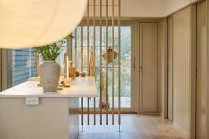 Interno cucina luminoso con isola e vaso, davanti a una parete divisoria WoodUpp: listelli verticali di legno chiaro ancorati a pavimento con piastre metalliche e barra orizzontale per appendere strofinacci e borse, creando separazione visiva verso la porta finestra, dall’atmosfera calda e minimalista.