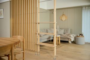 Divisorio modulare WoodUpp in legno chiaro con listelli verticali separa zona pranzo e soggiorno; collegata una struttura a scala WoodUpp con ripiani sottili, astine metalliche orizzontali e piedini regolabili, sullo sfondo divano grigio, plaid e piccolo tavolino d’appoggio.