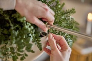 Mani che fissano un’asta tubolare sottile robusta in acciaio a un vaso sospeso bianco pieno di felci; si vede la clip di fissaggio grigia WoodUpp attaccata al bordo. Prodotti WoodUpp visibili: asta metallica per sospensione e clip/gancio adesivo per vaso.