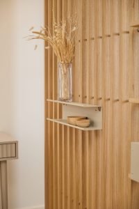 Pannello verticale WoodUpp a listelli in legno chiaro fissato a parete, con due mensole modulari WoodUpp color sabbia sospese; sulla mensola superiore un vaso di vetro con fiori secchi, sulla inferiore due ciotole in legno. Scena minimalista e naturale, con texture e finitura visibili.