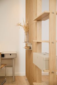Immagine WoodUpp: parete divisoria a doghe in rovere chiaro con scaffalatura modulare e mensole metalliche beige fissate ai profili, che reggono un vaso di vetro con graminacee essiccate e una piccola ciotola in legno; scrivania minimal e sedia in primo piano.