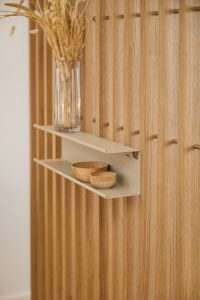 Parete WoodUpp composta da pannelli verticali in rovere a doghe chiaro, con mensola metallica beige WoodUpp montata a incastro. Sulla mensola coppia di ciotoline in legno e vaso di vetro con piante secche, illuminazione calda che esalta venature e texture naturali.