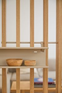 Divisorio WoodUpp composto da listelli verticali in rovere e mensola sospesa beige in metallo fissata con staffe; sulla mensola due ciotole artigianali in legno, una più grande e una più piccola, con venature visibili. Sfondo sfocato di un soggiorno con tavolino e libri.