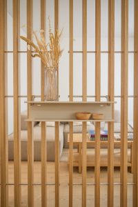 Divisorio a listelli in rovere WoodUpp con ripiano sospeso beige WoodUpp fissato da staffe metalliche; sul ripiano un vaso di vetro con fiori secchi e due ciotole in legno. Sullo sfondo soggiorno luminoso con divano e tavolino basso con libri.