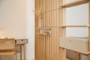 Parete divisoria WoodUpp a doghe verticali in legno chiaro con ripiani modulari beige e cassetto sospeso WoodUpp; sul ripiano due ciotole in legno e un vaso di vetro con fiori secchi. In primo piano scrivania chiara e sedia in paglia di Vienna.