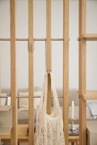 Divisorio modulare WoodUpp in listelli di rovere con barre metalliche orizzontali; si vedono una mensola a destra e due ganci in acciaio WoodUpp, uno dei quali regge una borsa rete bianca. Dettaglio minimalista, finitura naturale e accessori funzionali, perfetti per organizzare gli spazi con stile.