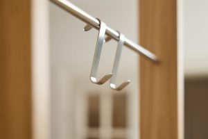 Immagine WoodUpp con due ganci in alluminio spazzolato che si agganciano a un'asta metallica: design sottile e piegato a U con estremità ricurve e marchio discreto sulla base. Appendiabiti minimal sospeso tra profili in legno, sfondo sfocato e atmosfera moderna.